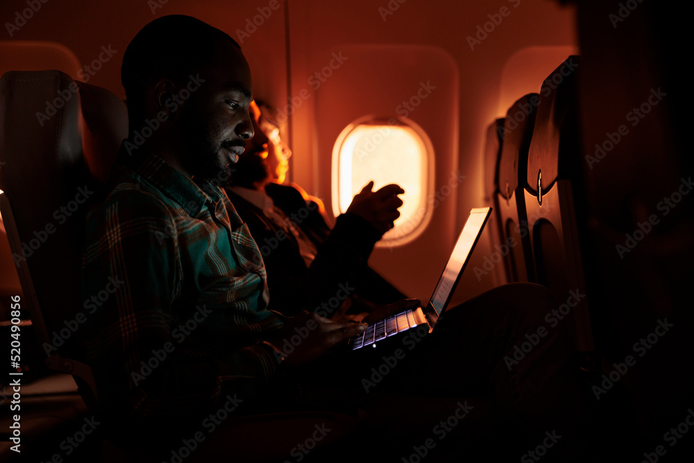 Male passenger browsing internet on laptop and flying abroad in airplane, using international airline to travel on business work trip. Young man working on computer and travelling during sunset.