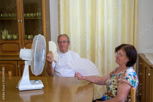  Image of an elderly couple at home wiping their sweat and trying to cool off with a fan and a fan. Reference to the great seasonal heat due to climate change
