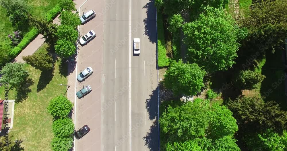 Following the white car going along the road surrounded by greenery. Residential area of a town from aerial perspective on sunny day. Top view. Following the white car going along the road surrounded by greenery. Residential area of a town from aerial perspective on sunny day. Top view.