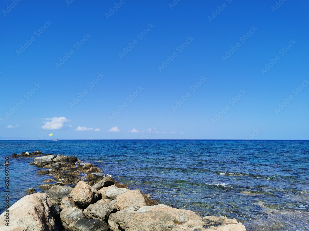 Coast of the Mediterranean Sea, waves, clear water, stone ridge against the blue sky with clouds boat with a yellow parachute.