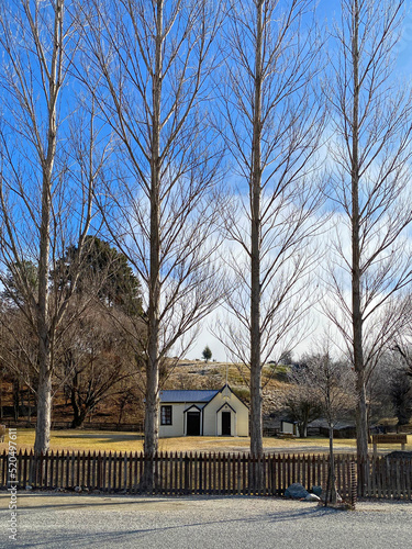 Cardrona village town hall with grass lawn and a picket fence viewed through trees.