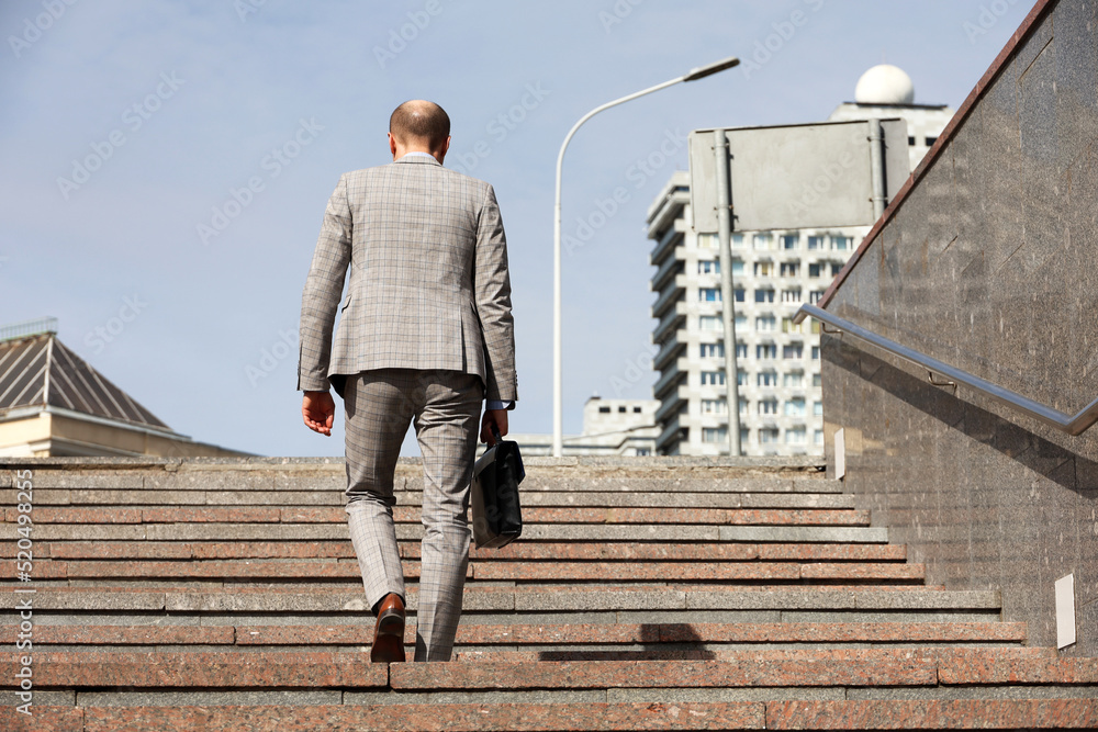 Man in a business suit with briefcase climbing stone stairs on sky and city buildings background. Concept of career, success, moving to the top, official or businessman Man in a business suit with briefcase climbing stone stairs on sky and city buildings background. Concept of career, success, moving to the top, official or businessman