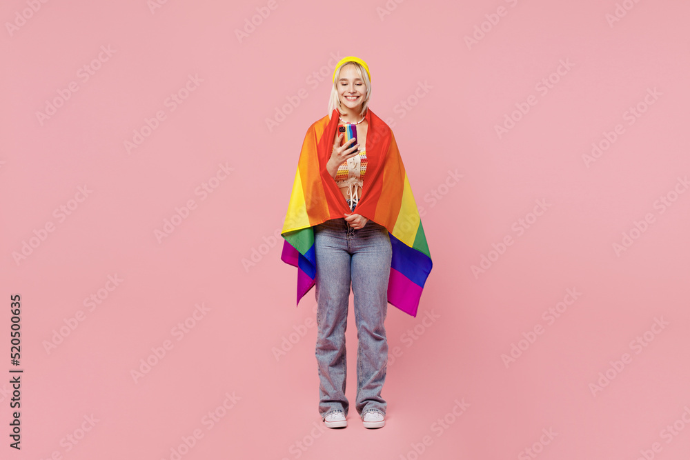 Full body young blond lesbian woman 20s wrapped in flag she wear colorful yellow hat hold in hand use mobile cell phone isolated on plain pastel light pink background. People lgbtq lifestyle concept. Full body young blond lesbian woman 20s wrapped in flag she wear colorful yellow hat hold in hand use mobile cell phone isolated on plain pastel light pink background. People lgbtq lifestyle concept.