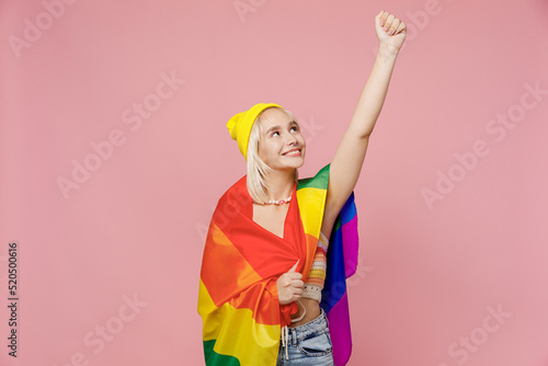 Young confident cool blond lesbian woman she wear yellow hat wrapped in striped flag do hero gesture clench fist isolated on plain pastel light pink background. People lgbtq lifestyle fashion concept.