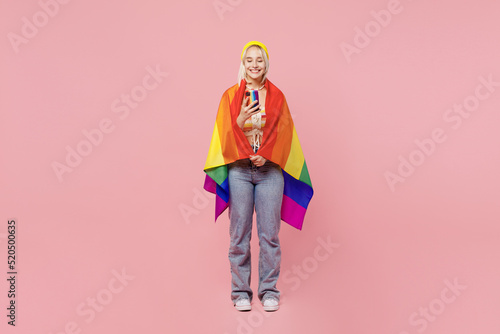 Full body young blond lesbian woman 20s wrapped in flag she wear colorful yellow hat hold in hand use mobile cell phone isolated on plain pastel light pink background. People lgbtq lifestyle concept.