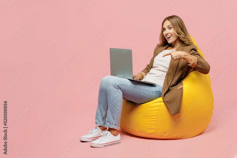 Full body young employee business woman 30s she in casual brown classic jacket sit in bag chair hold use work point finger on laptop pc computer isolated on plain pastel light pink background studio Full body young employee business woman 30s she in casual brown classic jacket sit in bag chair hold use work point finger on laptop pc computer isolated on plain pastel light pink background studio