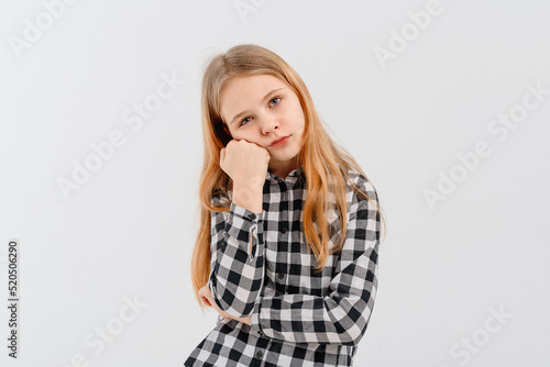 Cute teen girl looks bored or upset, sulks with head on fist, stands disappointed against white background, wears casual clothes