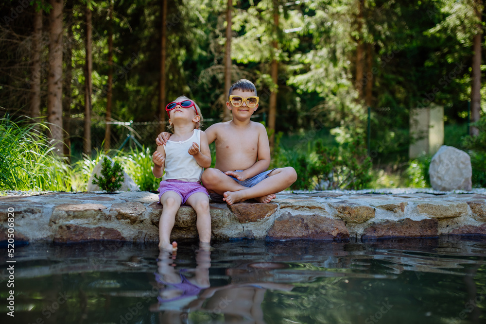 Fototapeta premium Cute girl and boy sitting together on the footpath by the lake with sun glasses, dangle their feet in the water, summer vacation concept.