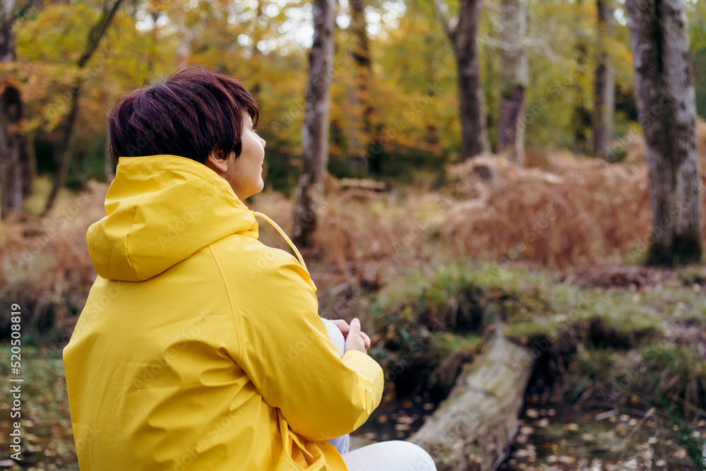 Back view woman in bright yellow raincoat sitting on a fallen tree, looking at the forest and enjoying the moment. Feeling harmony, reunion with nature in autumn. Relaxing, personal fulfillment Back view woman in bright yellow raincoat sitting on a fallen tree, looking at the forest and enjoying the moment. Feeling harmony, reunion with nature in autumn. Relaxing, personal fulfillment