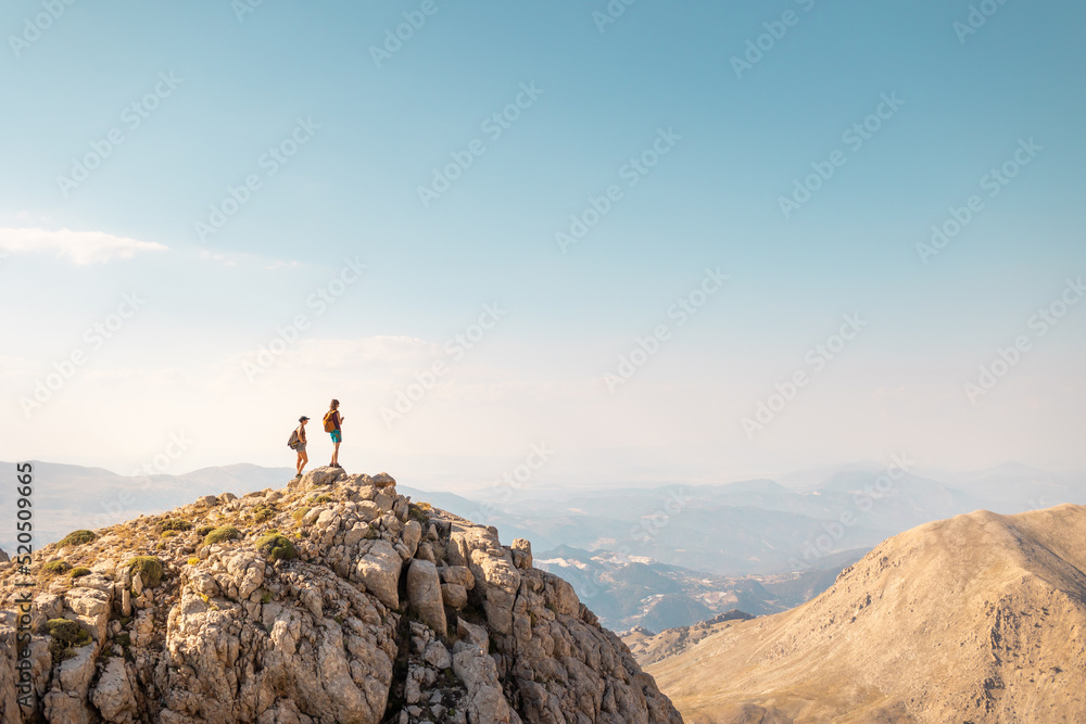 two girls with backpacks stand on top of a mountain.