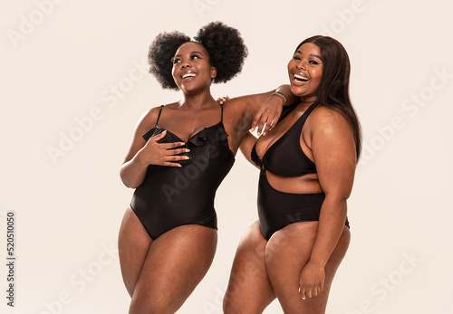 Two smiling, beautiful female models in black fashionable swimsuit posing together in studio, having fun together. Body positive concept. Plus size women.