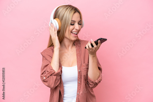 Pretty blonde woman isolated on pink background listening music with a mobile and singing
