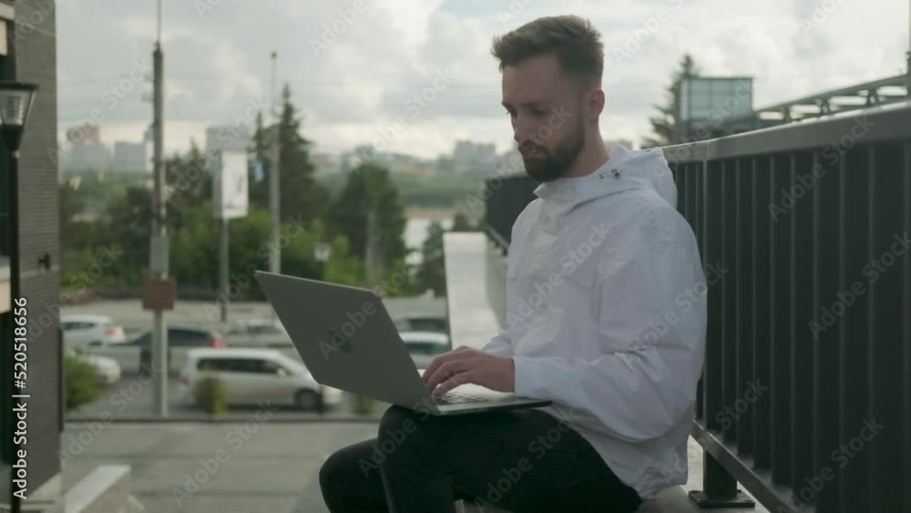 Young successful guy with laptop is sitting on stone bench working on computer. person outside is working remotely on street background of city streets. student is resting and using laptop outside Young successful guy with laptop is sitting on stone bench working on computer. person outside is working remotely on street background of city streets. student is resting and using laptop outside