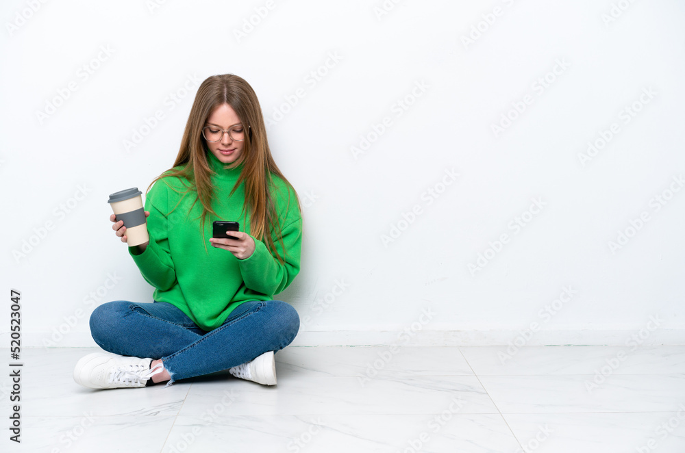 Young caucasian woman sitting on the floor isolated on white background holding coffee to take away and a mobile Young caucasian woman sitting on the floor isolated on white background holding coffee to take away and a mobile