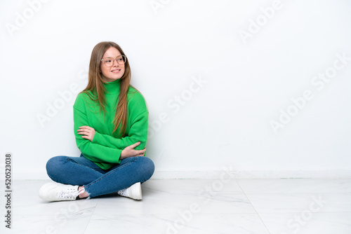 Young caucasian woman sitting on the floor isolated on white background with arms crossed and happy