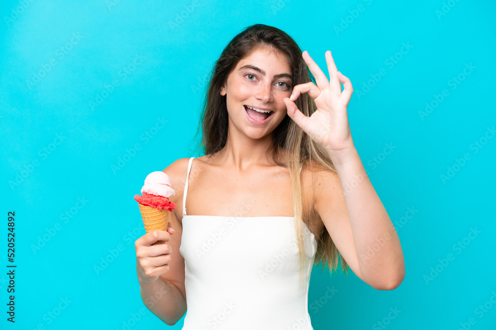 Young woman in swimsuit holding an ice cream isolated on blue background showing ok sign with fingers Young woman in swimsuit holding an ice cream isolated on blue background showing ok sign with fingers