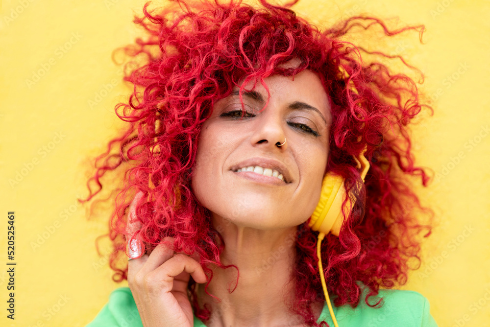 close-up of a smiling young latin woman with red afro hair wearing a blue t-shirt enjoys listening to music holding headphones with one hand on a yellow background looking at the camera close-up of a smiling young latin woman with red afro hair wearing a blue t-shirt enjoys listening to music holding headphones with one hand on a yellow background looking at the camera