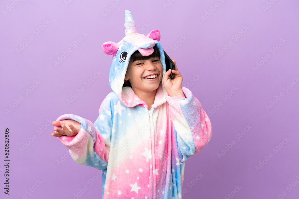 Little kid wearing a unicorn pajama isolated on purple background keeping a conversation with the mobile phone with someone