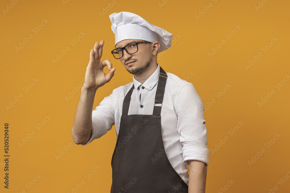 a male cook in glasses, a black apron and a beret with folds shows an OK sign, stands on a yellow background a male cook in glasses, a black apron and a beret with folds shows an OK sign, stands on a yellow background