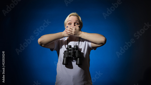 A blonde woman with a blue-yellow Ukrainian flag on her cheek with binoculars around her neck, covered her mouth with her hands on a blue background. The concept says nothing.
