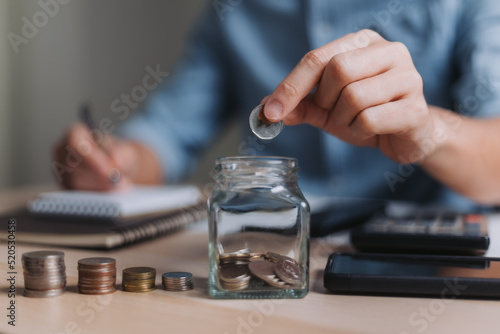 Businessman holding a coin in a glass with using a pen to write a notebook and using a calculator to calculate savings ideas for financial accounting.
