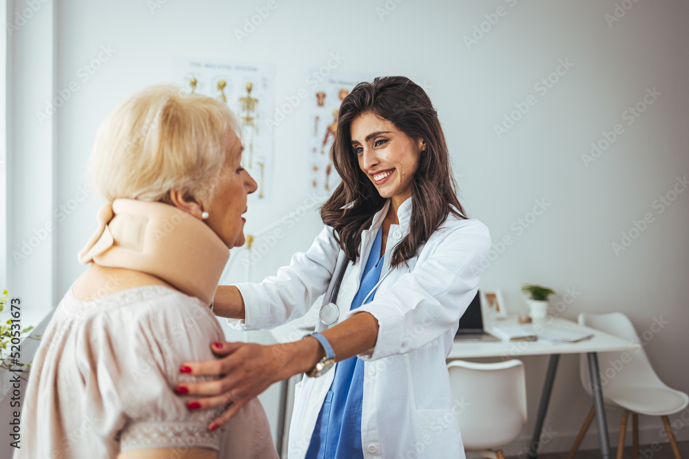 Fototapeta premium Female doctor putting neck orthopaedic collar on adult injured woman. Picture of senior woman having a visit at female doctor's office. Doctor helping a senior woman with her neck injury