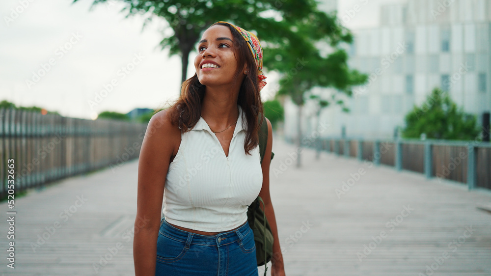 Сute tanned woman with long brown hair in white top and yellow bandana with backpack on her shoulder and looks around the modern city.