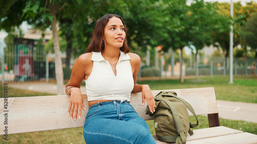 Naklejka premium Lovely tanned woman with long brown hair wearing white top sitting relaxed on park bench looking away. Life style concept. Slow motion.