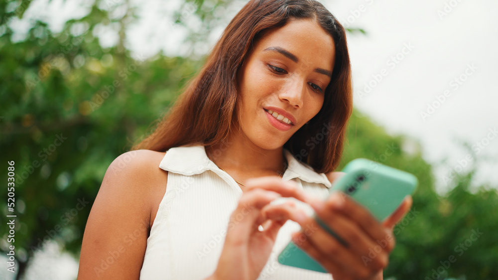 Clouse-up, beautiful girl with long dark hair wearing white top sits on bench and uses mobile phone. Young smiling woman writing in social networks on cellphone.