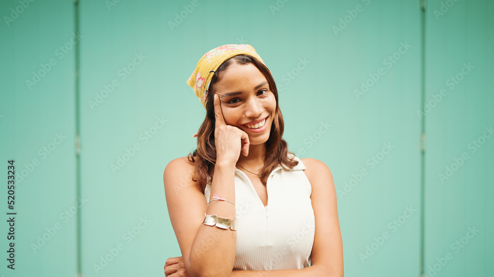 Beautiful girl with tanned skin, brown long hair and bandanna on her head poses for the camera. Smiling young woman putting one finger to the cheek on blue background Beautiful girl with tanned skin, brown long hair and bandanna on her head poses for the camera. Smiling young woman putting one finger to the cheek on blue background