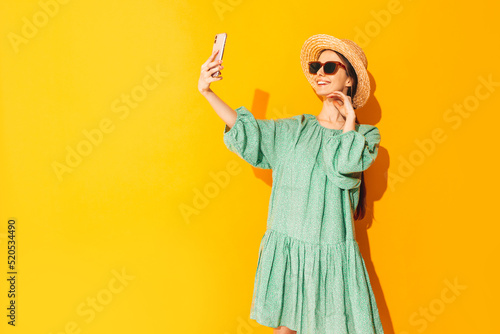 Young beautiful smiling female in trendy summer green dress. Sexy carefree woman posing near yellow wall in studio. Positive model having fun. Cheerful and happy. In hat. Isolated. Taking selfie