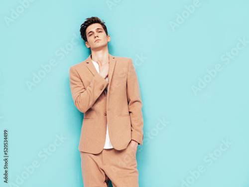 Portrait of handsome confident stylish hipster lambersexual model. Sexy modern man dressed in elegant beige suit. Fashion male posing in studio near blue wall