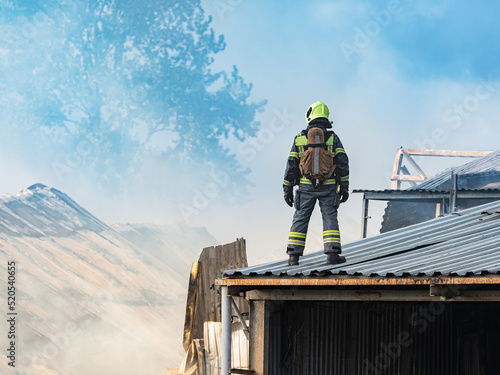 Firefighter on roof of building. Emergency Specialist. Man in firefighter uniform backs camera. Roof house ha covered in smoke. Rescuer examines source ignition. Concept emergency in industrial area © Grispb