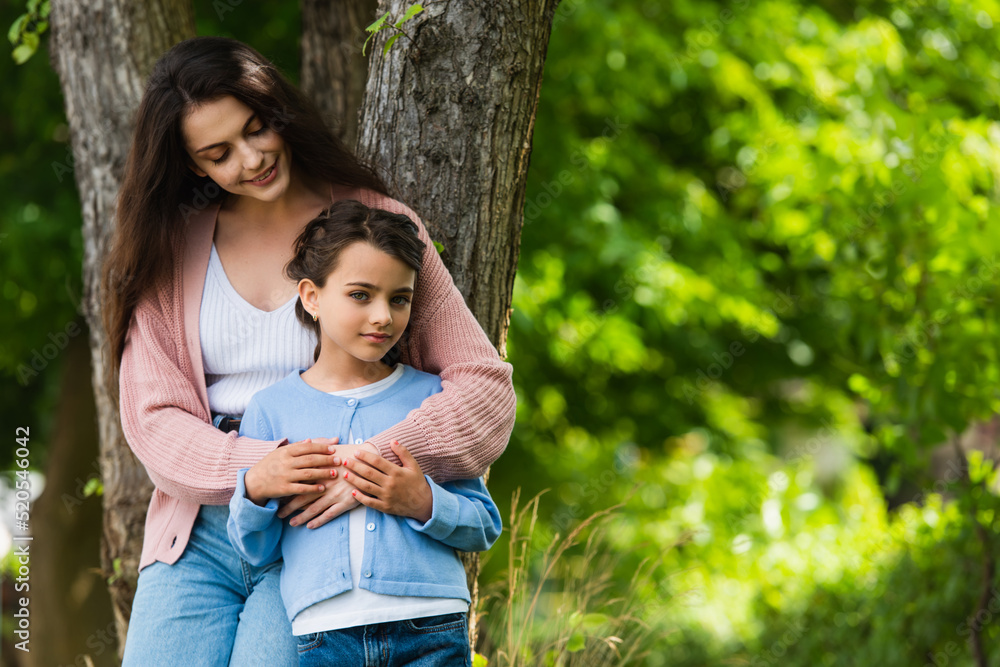 happy young woman hugging daughter near trees in park happy young woman hugging daughter near trees in park