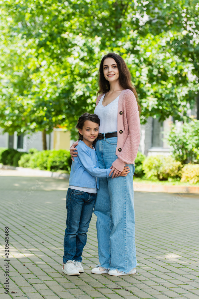 full length of mom and daughter in jeans embracing and smiling at camera on city street