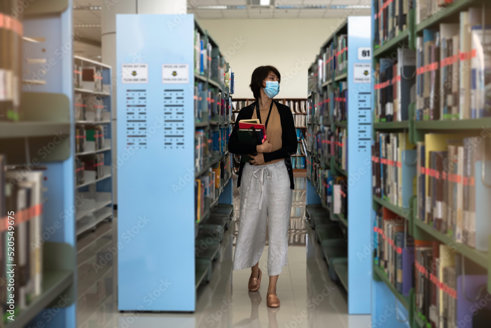 Woman with face mask hold hard cover books in hands,walking between shelf, Woman with face mask hold hard cover books in hands,walking between shelf,