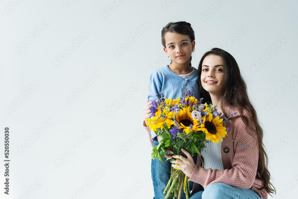 woman and girl smiling at camera near bouquet of blue and yellow flowers isolated on grey woman and girl smiling at camera near bouquet of blue and yellow flowers isolated on grey