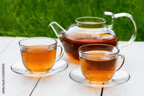 Tea in glass teapot and two cups on a wooden white background background against the background of green grass. Teapot with green or black tea. Tea time