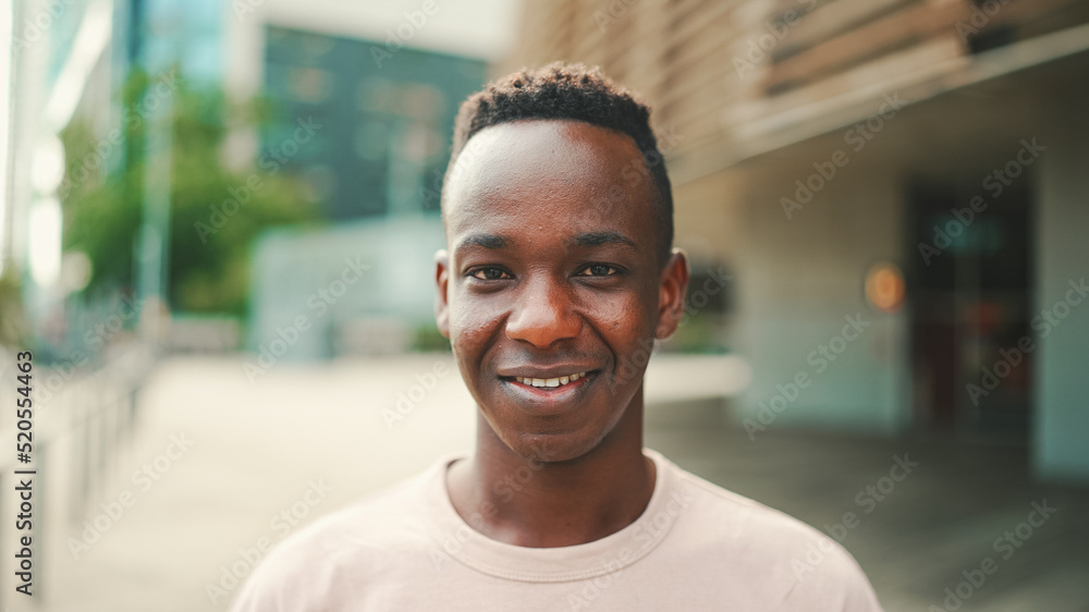Clouse up, young african student stands outside of the university, raises his head, looks at the camera, smiles Clouse up, young african student stands outside of the university, raises his head, looks at the camera, smiles