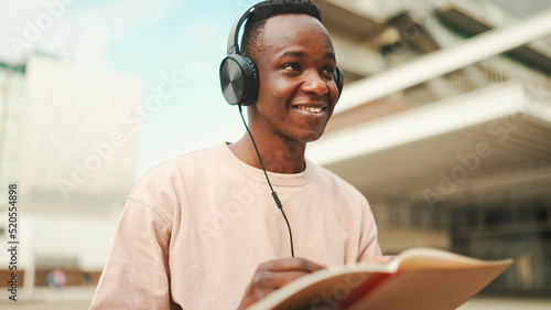 Young african student sitting outside of university and taking notes while study online with headphones © Andrii Nekrasov