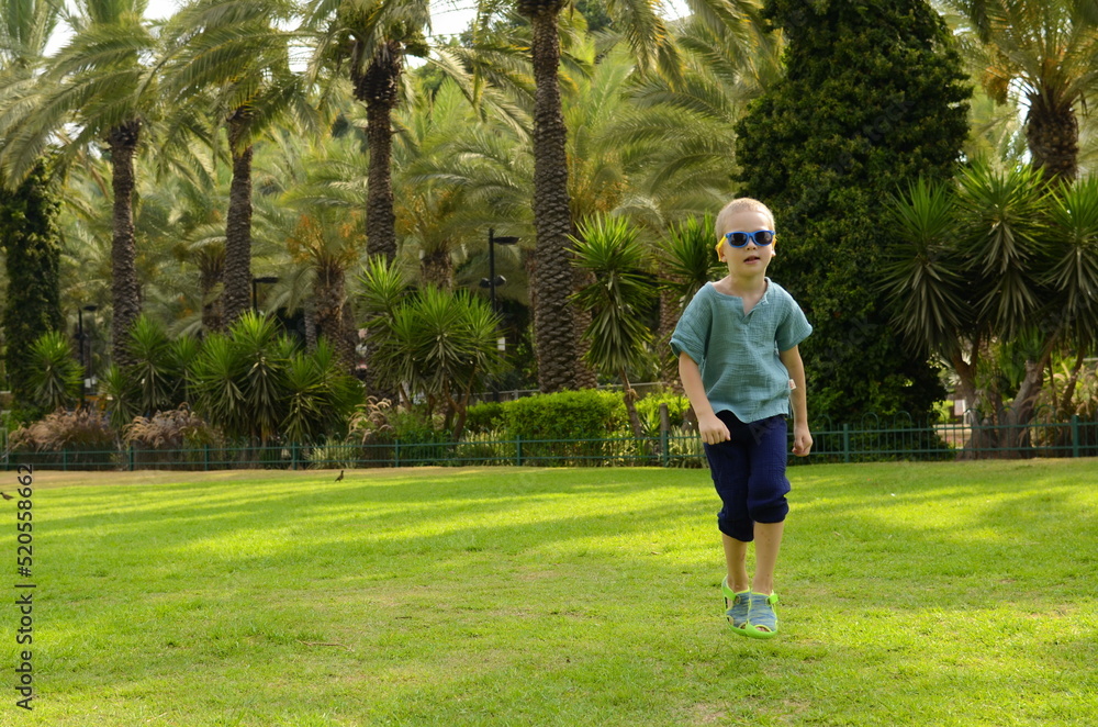 The boy jumps for joy on a green field. A child in sunglasses on a background of palm trees. Concept: holidays, travel, exotic destinations, tropical countries.