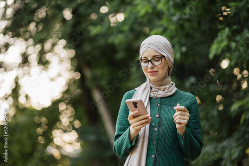 Portrait of beautiful muslim female in casual clothes watching online story on mobile phone, connected with wireless internet. Young cute girl with hijab listen music from smartphone walking on street © sofiko14