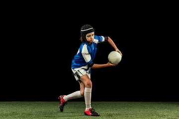 Dynamic portrait of school age boy, junior male rugby player practicing rugby football isolated on dark background with grass floooring.