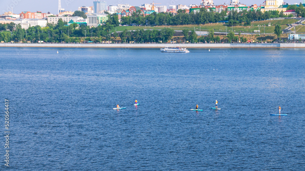 Obraz premium Active recreation on the board on the big river. Tourism on sap boards. Outdoor water sports. Surfers, standing on a board, ride the waves. SUP surfing training. View of the city embankment