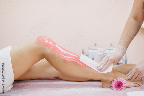 A master applies pink depilatory wax to a young woman's leg for hair removal. Strip for depilation. Depilation with wax. Beauty concept. Place for text.