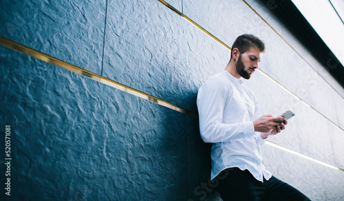 Concentrated guy using smartphone near wall of modern building