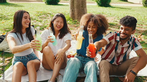 Happy, smiling multiethnic young people at picnic on summer day outdoors. Group of friends having fun with drinks, raising toasts while relaxing in the park at picnic