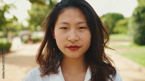 Close-up young Asian smiling woman with long brown hair wearing white t-shirt posing for the camera in the park . The girl opens her eyes and smiles at the camera