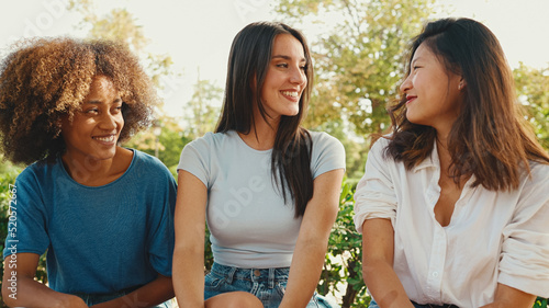 Happy multiethnic young women talking while sitting on park bench on summer day outdoors. Group of girls talking and laughing merrily in city park