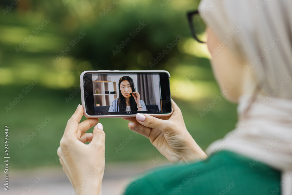 Smart phone screen view. Close up of unrecognizable female, sitting at green park and holding smart phone while talking with her office friend colleague, young Asian lady in headset.